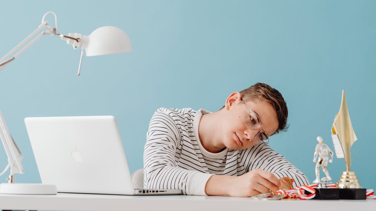 Teenager with eyeglasses contemplating medals at desk with laptop and trophies.
